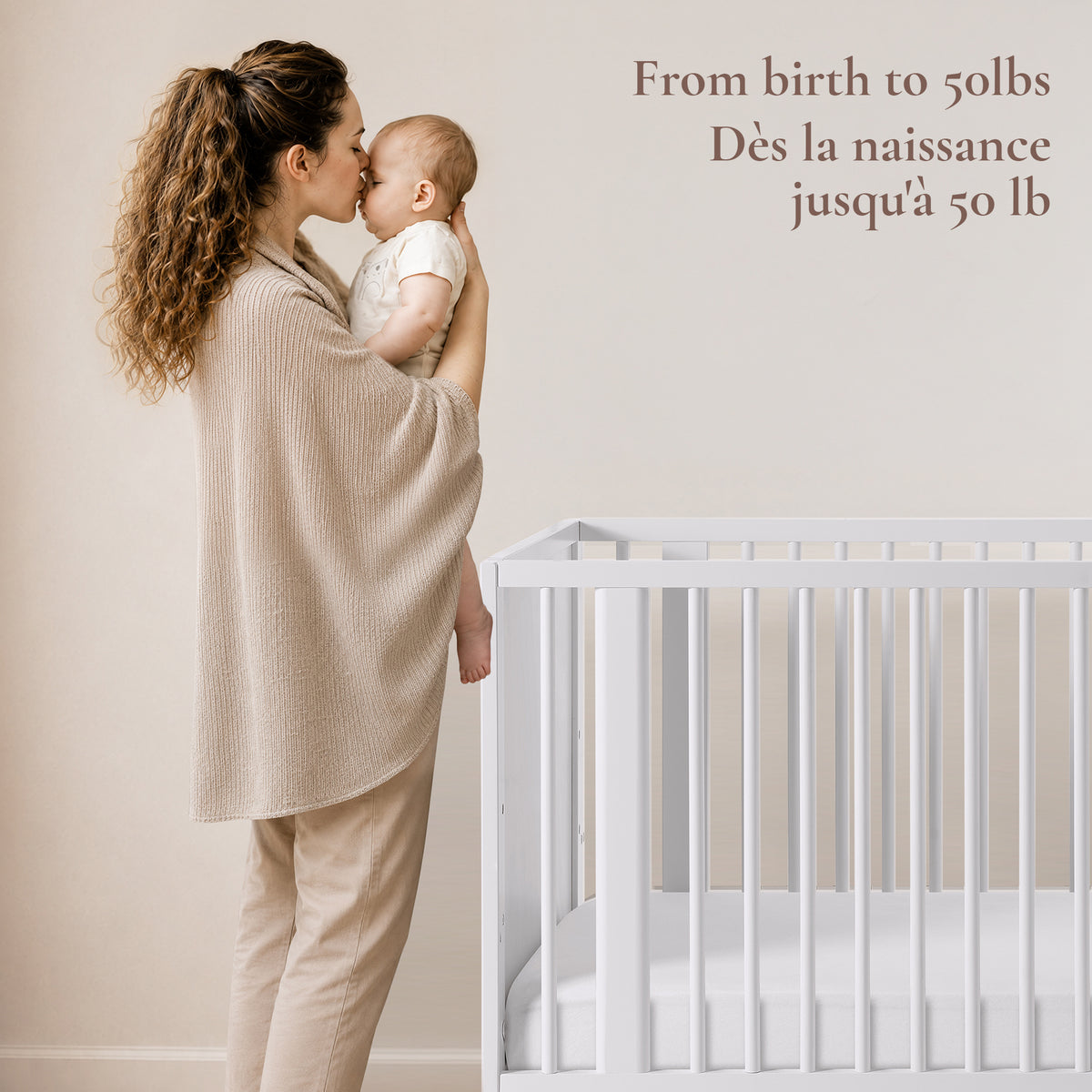 Lifestyle shot of a mother holding her baby beside the Roo 4-in-1 crib, highlighting safe use from birth up to 50 lbs.